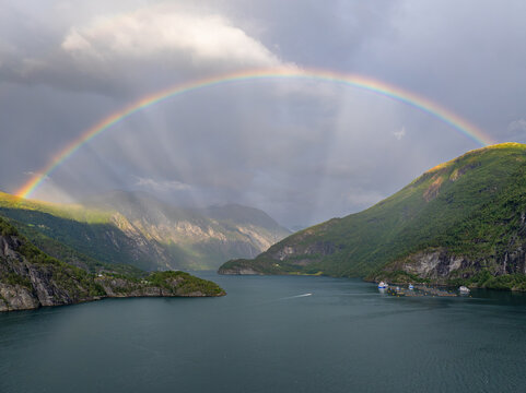 Aerial view of a fish farm situated in a deep fjord surrounded by steep mountains under a dramatic sky with a vibrant rainbow in Fjord, More og Romsdal, Norway.