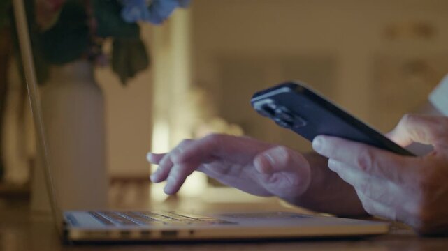 Detailed shot of a man's hands multitasking at night, using a smartphone and typing on a laptop computer, representing the concept of modern digital workflow and constant connectivity