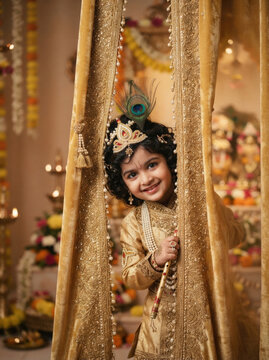 Little boy dressed as lord krishna holding bamboo flute peeking through golden silk curtains celebrating janmashtami festival event.