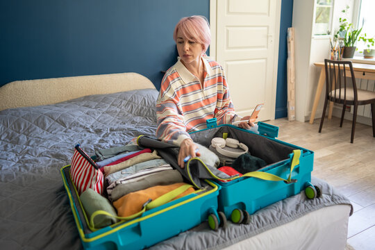 Woman traveler preparing luggage for trip and revising items in suitcase with checklist in notepad, focused female checking clothes and accessories in open travel case while sitting on bed in bedroom.