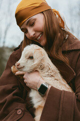 Obraz premium Joyful woman in autumn coat holds a young goat kid close, sharing affectionate moment outdoors, warm scarf, smile, gentle interaction, rustic mood, lifestyle scene, candid portrait, natural light,