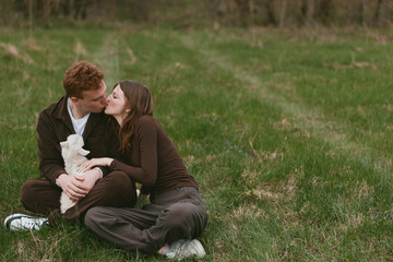 Obraz premium Loving couple outdoors kiss while sitting on a grassy field, holding a small white puppy, casual clothes, relaxed mood, candid lifestyle moment celebrating affection and companionship in nature.