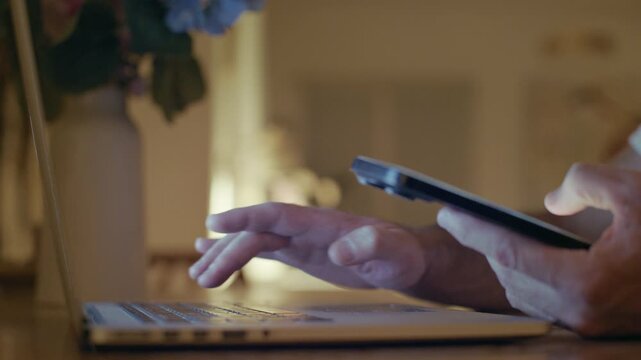 Close up of a person's hands multitasking at home, using the trackpad on a laptop computer with one hand while simultaneously scrolling on a smartphone with the other in a cozy interior