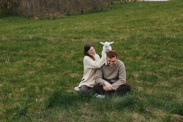Obraz premium Happy couple sits on a grassy field with a playful goat kid between them, sharing a gentle moment in cozy sweaters, natural light, candid lifestyle portrait outdoors, loving interaction and rural