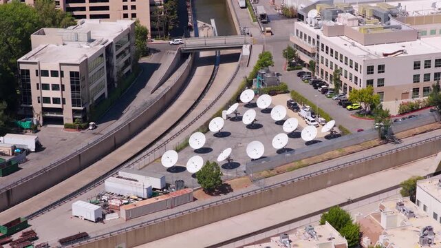 Aerial view of white satellite dish antennas array at television broadcast facility. Media studio lot with office buildings and concrete river channel in Los Angeles on sunny summer day.
