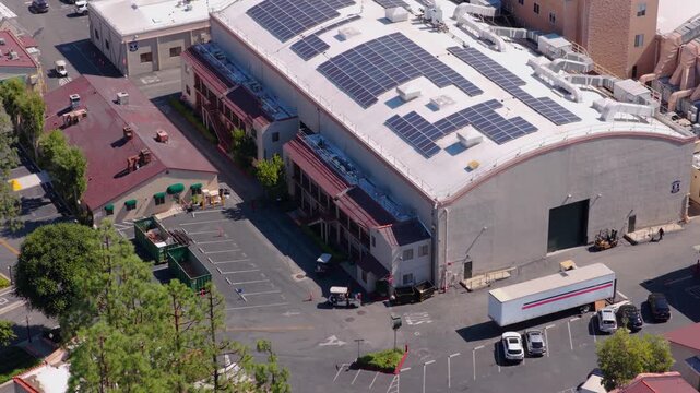 Aerial view of large sound stage building with solar panels on curved white roof. Film studio backlot with trucks golf carts and parking. Sunny summer day in California. Clean energy production.