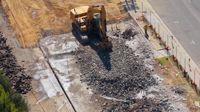 Aerial view of yellow excavator breaking concrete rubble on urban demolition site. Construction worker in safety vest watching heavy machinery work. Sunny day industrial teardown from drone.