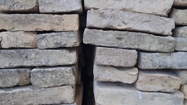 Detailed camera zoom on stacked ancient clay bricks and weathered stone slabs stored outdoors, highlighting raw textures and traditional building materials.