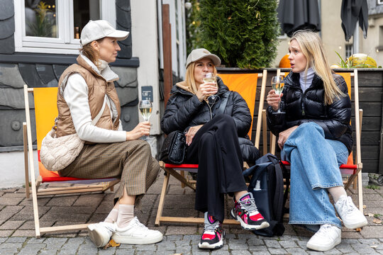 Three women sit outdoors with wine, sharing gossip and lively discussion during city break in Germany. Candid lifestyle scene shows friendship, conversation, leisure, travel and social connection