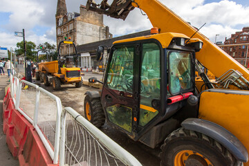 A busy road construction site on a city street under sky with scattered clouds. A yellow telescopic handler (telehandler) on a street construction site. Area is separated from the pedestrian walkway.  © Jacek