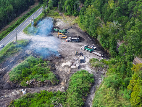 Aerial view of a construction site with heavy machinery, dump trucks, and cleared land surrounded by dense forest near a road in Wilmington, North Carolina, United States.