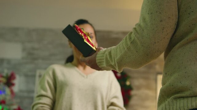 Man keeps a wrapped present concealed behind his back as he approaches a woman in a festive living room, building anticipation for a Christmas gift reveal, highlighting Christmas surprise.