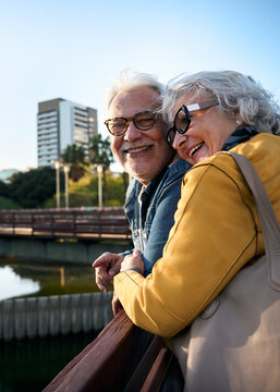 Vertical. Smiling senior couple leaning on a railing in a city park, enjoying the view of a pond and cityscape, embracing their golden years together