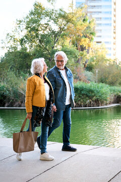 Senior couple walking and talking beside a lake in a green city park at sunset. Candid outdoor scene conveying companionship, active aging, affection and a peaceful shared lifestyle.