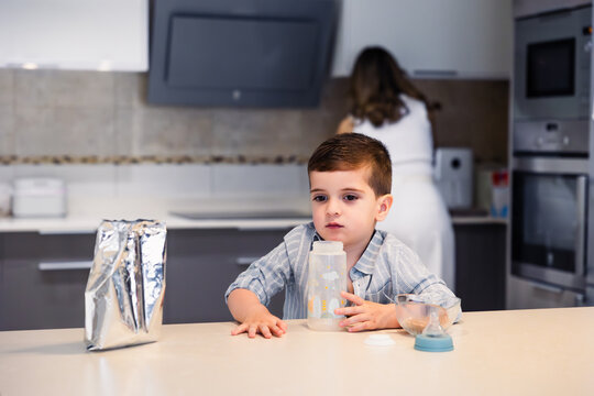 Toddler sitting at kitchen table holding bottle watching mother