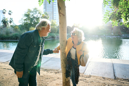 Happy senior couple playing around a tree and laughing beside a lake in a sunlit park. Candid outdoor scene conveying affection, humor, companionship and joyful active aging.