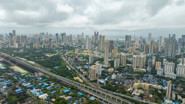 Aerial view of the dense Mumbai cityscape featuring modern skyscrapers, high-rise buildings, a sprawling railway line, and green patches under a cloudy sky in Mumbai, Maharashtra, India.