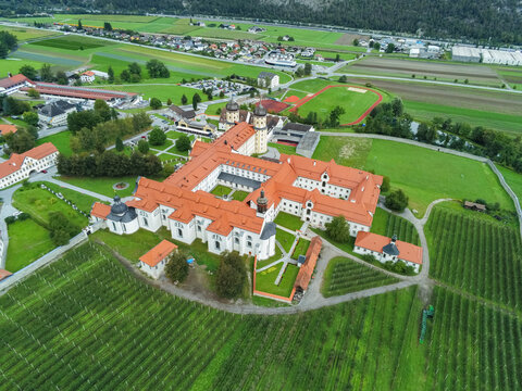 Aerial view of Stift Stams, a historic Cistercian monastery with red-tiled roofs and white walls, surrounded by lush green fields and vineyards in Stams, Tirol, Austria.