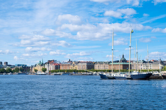 Classic two-masted sailing boat anchored in the harbor along Strandvagen boulevard with colorful historic buildings, blue sea, and cloudy sky in Stockholm Sweden