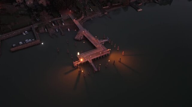 Aerial view of the illuminated pier at Isola Maggiore extending into Lake Trasimeno at dusk with warm lights reflecting on the water in Tuoro sul Trasimeno, Umbria, Italy.