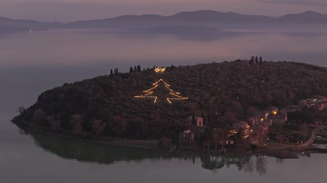 Aerial view of the illuminated Christmas tree light display on the hillside of Isola Maggiore at dusk in Tuoro sul Trasimeno, Umbria, Italy.