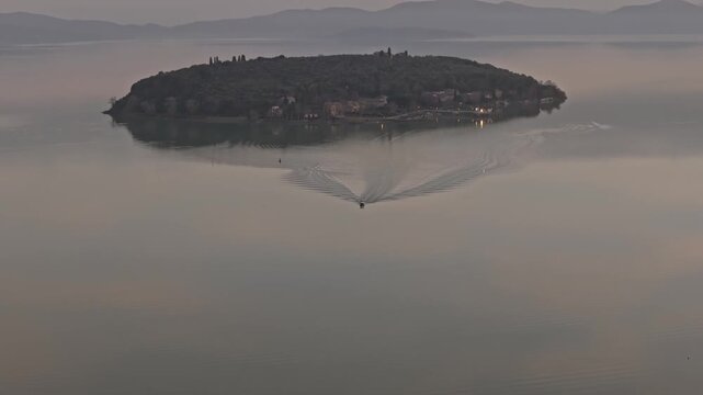 Aerial view of Isola Maggiore on Lake Trasimeno with a small boat leaving a wake in the calm water in Tuoro sul Trasimeno, Umbria, Italy.