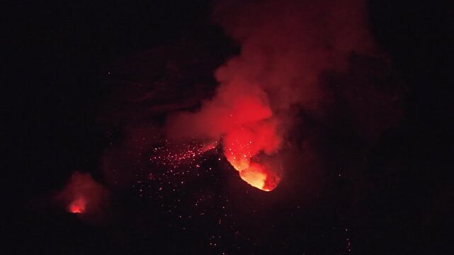 Aerial view of Stromboli volcano erupting at night with glowing lava and volcanic ash clouds rising into the dark sky in Stromboli, Sicily, Italy.