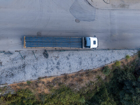 Aerial view of a white flatbed truck transporting a load of long metal pipes along a paved road next to a stone-paved area and greenery in Istanbul, Istanbul, Turkiye.