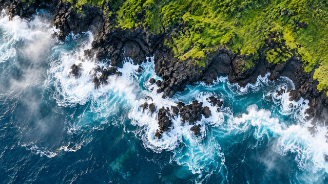 Top-down aerial view of powerful turquoise waves exploding into white foam against jagged black volcanic rocks, vivid green coastal moss and a rugged remote shoreline.