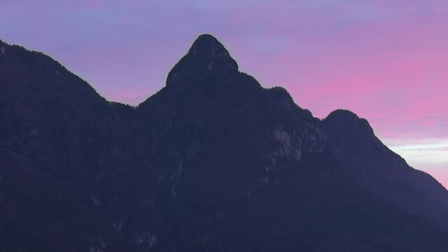 Aerial view of rugged mountain peaks silhouetted against a vibrant purple and pink sunset sky in Longarone, Veneto, Italy.