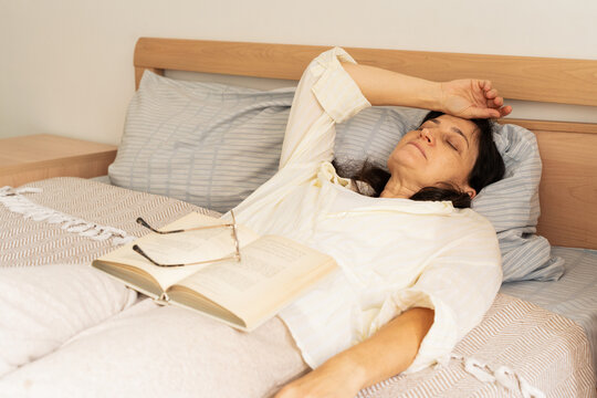 Mature woman enjoys leisurely morning at home, resting in bed with book and glasses. A measured pace of life, mindful rest, self-care, and calm daily routine. Philosophy of slow living