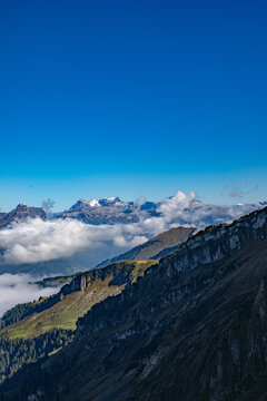 Aerial view of the Swiss Alps with snow-capped peaks, rolling green slopes, and a sea of clouds under a clear blue sky in Airolo, Ticino, Switzerland.