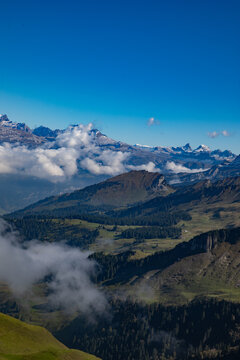 Aerial view of the Swiss Alps with snow-capped peaks, rolling green valleys, dense pine forests, and clouds drifting over the landscape in Airolo, Ticino, Switzerland.