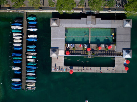 Aerial view of the Frauenbad Stadthausquai public bath with people swimming, red umbrellas on the wooden deck, and a pier with moored boats in the Limmat river in Zurich, Zurich, Switzerland.
