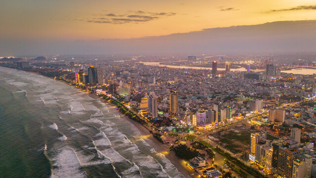 Aerial view of the coastal city of Da Nang at sunset, featuring high-rise hotels, sandy beaches, and the Han River flowing through the urban landscape in Da Nang, Vietnam.