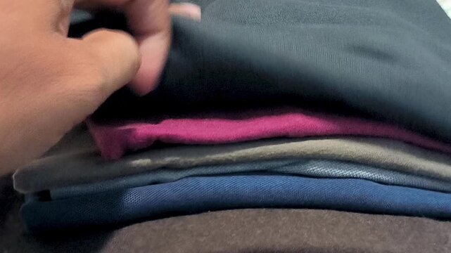 A close-up shot of a hand flipping through a stack of neatly folded colorful t-shirts and garments