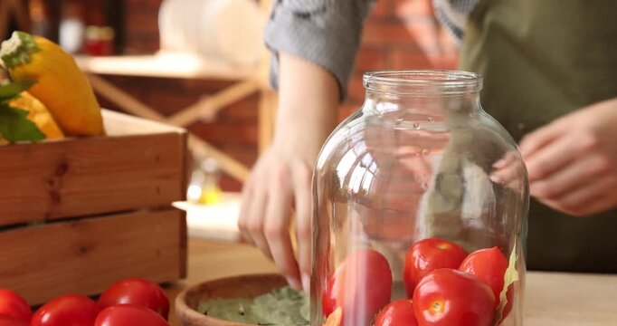 Woman putting bay leaves into pickling jar with tomatoes at table indoors, closeup