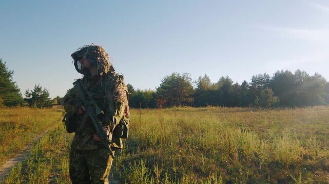 Soldier in ghillie suit carrying rifle walks through grassy field during sunny day.