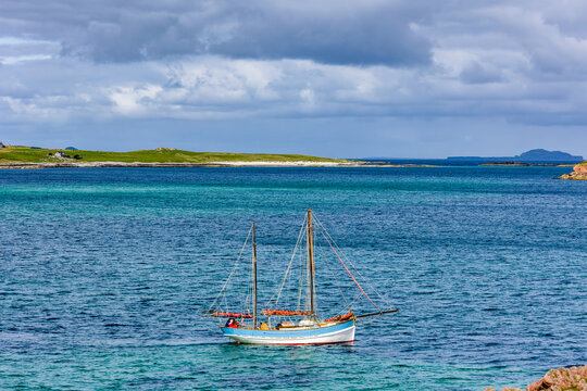 Traditional two-masted wooden boat floating on turquoise coastal water, low green hills and sandy shore of Iona visible across sea channel
