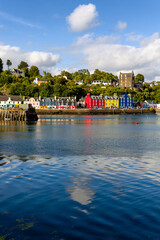 Fototapeta premium Colorful painted houses along Tobermory harbor waterfront, Scotland, calm blue sea water reflecting bright facades under wooded hillside