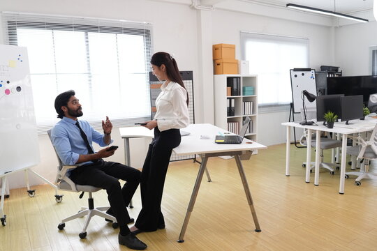 Candid workplace banter with young Indian businessman and Asian businesswoman at desk area in front of whiteboard, soft daylight, everyday business life, corporate lifestyle