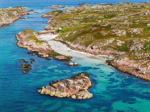 Clear turquoise sea around pink granite rocks and sandy inlet, green moorland coastal landscape on Scottish island in summer, aerial view