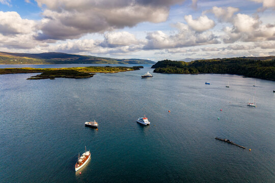 High angle aerial perspective of yachts and boats floating on blue coastal waters, wooded shoreline and low green hills stretch into distance