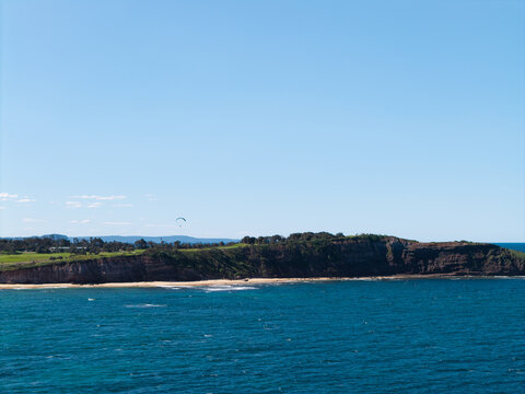 Aerial view of the Northern Beaches coastline featuring rugged cliffs, sandy beach, blue ocean, and a paraglider in the sky in Northern Beaches Council, New South Wales, Australia.