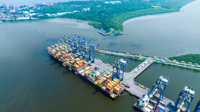 Aerial view of the Jawaharlal Nehru Port Authority container terminal with a large cargo ship, gantry cranes, and trucks on the access road in Juna Sheva, Maharashtra, India.