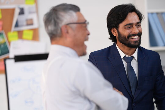 Close-up of senior Asian businessman and Indian businessman, team celebration in a modern office, near glass sticky-note, authentic candid moment, everyday business life