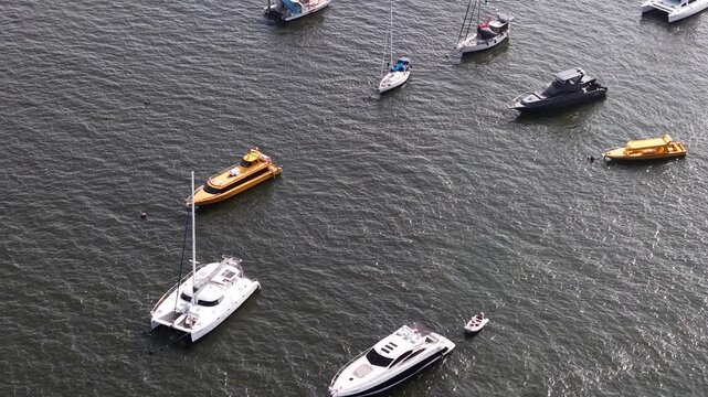Luxury yachts, catamarans, and small boats anchored in the rippling harbor waters evoke an atmosphere of leisure, summer travel, and a nautical lifestyle all framed within a powerful composition