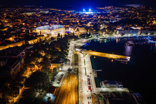 Aerial view of the illuminated Pula Arena at night, overlooking the historic city center, the bustling harbor with docked boats, and the coastal promenade in Pula, Istria County, Croatia.