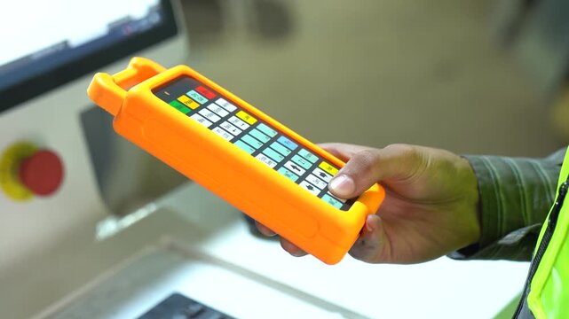 Industrial worker hand operating orange cnc remote controller at modern manufacturing workshop.