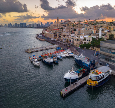 Aerial view of the Old port of Jaffa with fishing boats docked at the pier, historic stone buildings, and the Tel Aviv skyline at sunset in Tel Aviv-Yafo, Tel Aviv District, Israel.